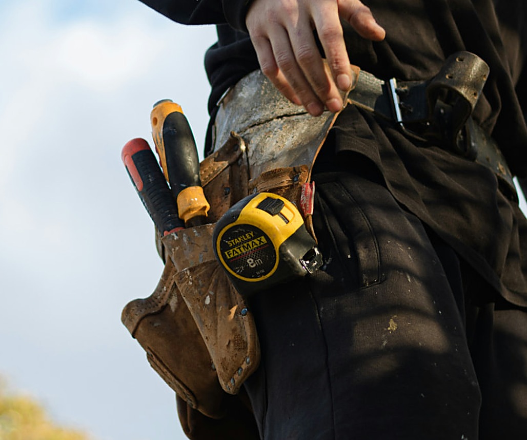 A handyman’s tool belt holding a tape measure and screwdrivers, resting on framing lumber at a job site